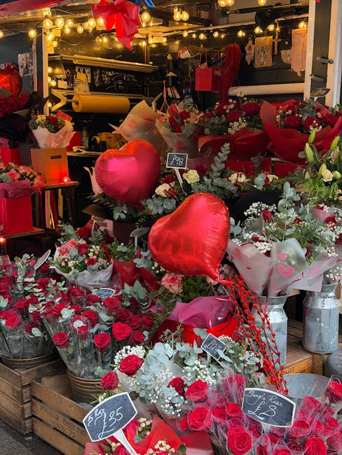 A vibrant flower display featuring red roses, white baby's breath, and green foliage arranged in various bouquets and wrapped in pink, red, and white paper. Large red helium balloons shaped like heart