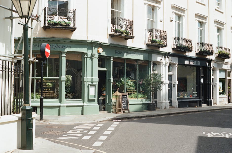 Exterior view of a quaint flower shop on Balham High Road, with large glass windows framed by a pastel green facade. The shop displays an array of fresh flowers and greenery inside, visible through th