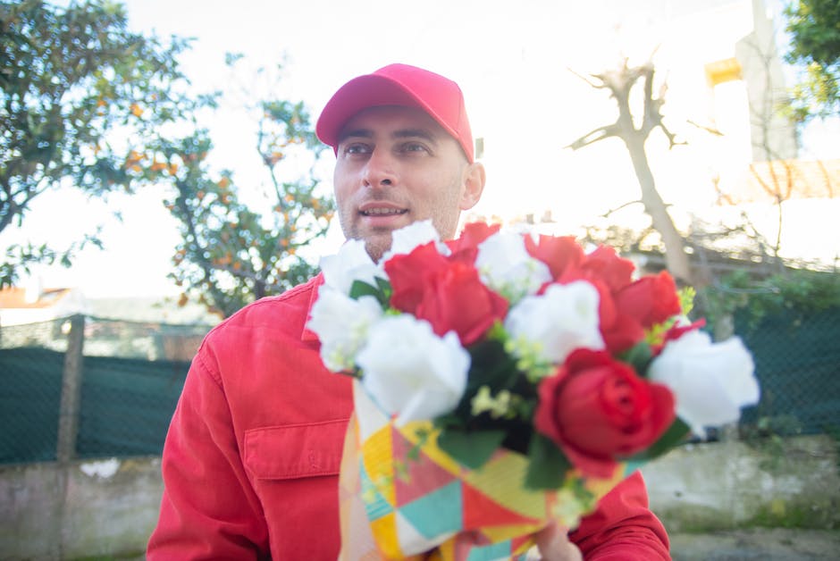 A delivery person in a red cap and matching red jacket holding a vibrant floral arrangement in a colorful patterned paper wrapping. The bouquet features an assortment of fresh flowers including red ro