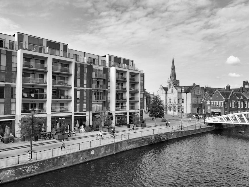 A modern multi-story residential building with large glass balconies facing a riverside promenade, where several pedestrians and cyclists are visible. In the background, a historic church with a tall 