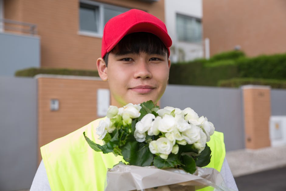 A young delivery person, wearing a red cap and a high-visibility yellow vest, stands outdoors in front of modern residential buildings holding a bouquet of fresh white flowers wrapped in white paper. 