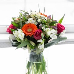 This sophisticated bouquet, crafted by skilled hands familiar with Balham's leafy avenues, showcases a masterful harmony of colour and shape. Soft peach gerbera daisies seem to glow with the warmth of a new day, their delicate petals fanning out around bold, fiery orange companions, reminiscent of sunrise skies over Bedford Hill. Deep, fuchsia roses add a sumptuous sense of romance and draw the gaze inward, while clusters of pristine white alstroemeria and miniature white roses infuse the arrangement with lightness and cheerful contrast. The bouquet's abundant green foliage is highlighted by wide, elegantly curved aspidistra leaves, enfolding the flowers in a protective arc that echoes the inviting gardens of Balham's classic Victorian homes. Resting in a tall, slender glass vase, the bouquet's lively green stems are on full display, reinforcing the feeling of freshness and abundance. The softly illuminated, bright background allows each flower to pop with colour and grace-a perfect centrepiece for gatherings, thoughtful gestures, or offering comfort and joy to a neighbour. Every detail reflects artisanal care and an understanding of local style, making it a true embodiment of Balham's community spirit.